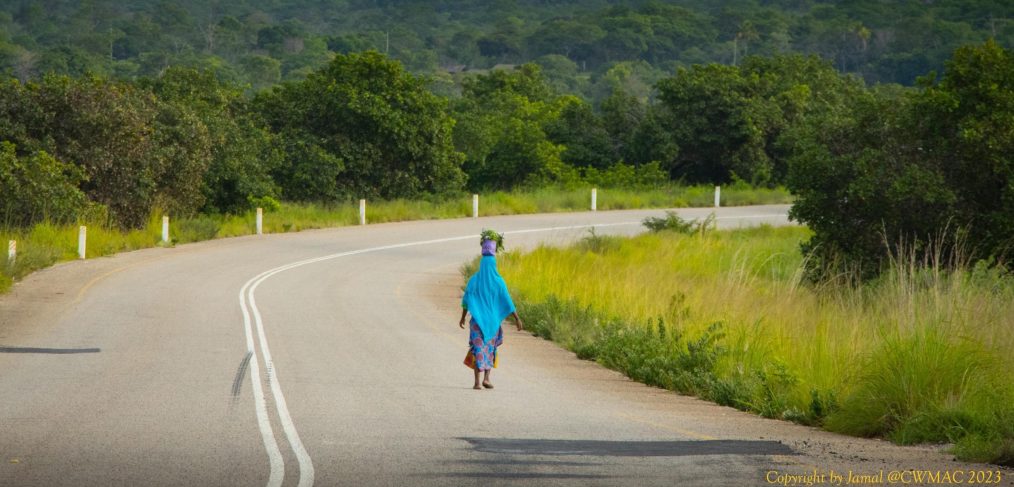 I woman with a basket on her head walks along a tarred road.