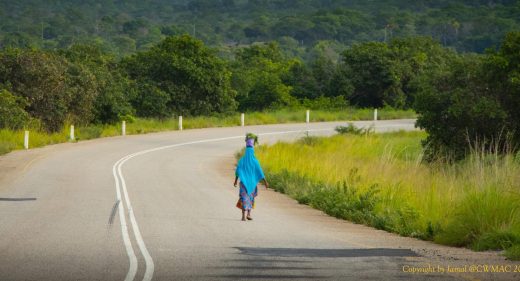 I woman with a basket on her head walks along a tarred road.