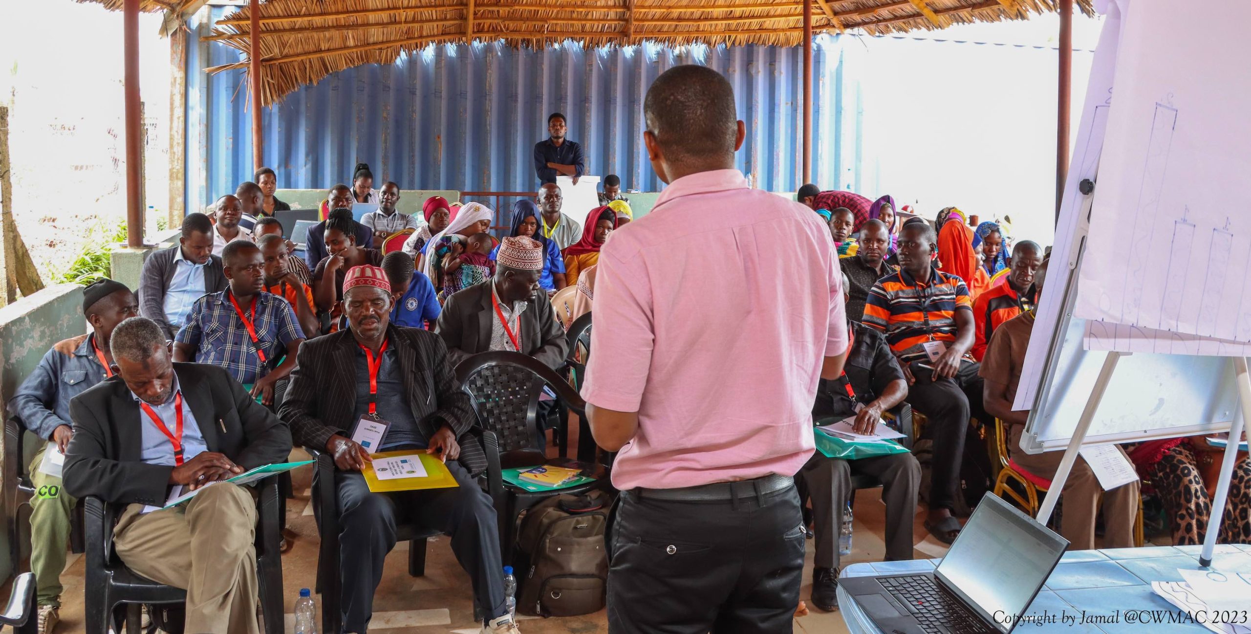A group of participants looking towards the camera and lecturer in an open-sided room.