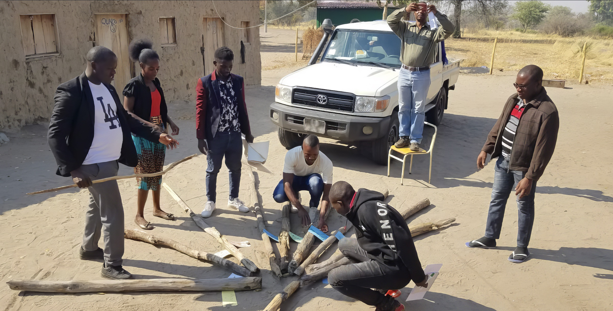Seven people examine pieces of paper attached to a pile of logs.