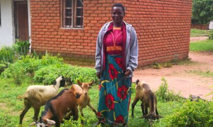 A smiling woman outside a red brick house with 4 goats.