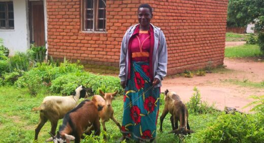 A smiling woman outside a red brick house with 4 goats.