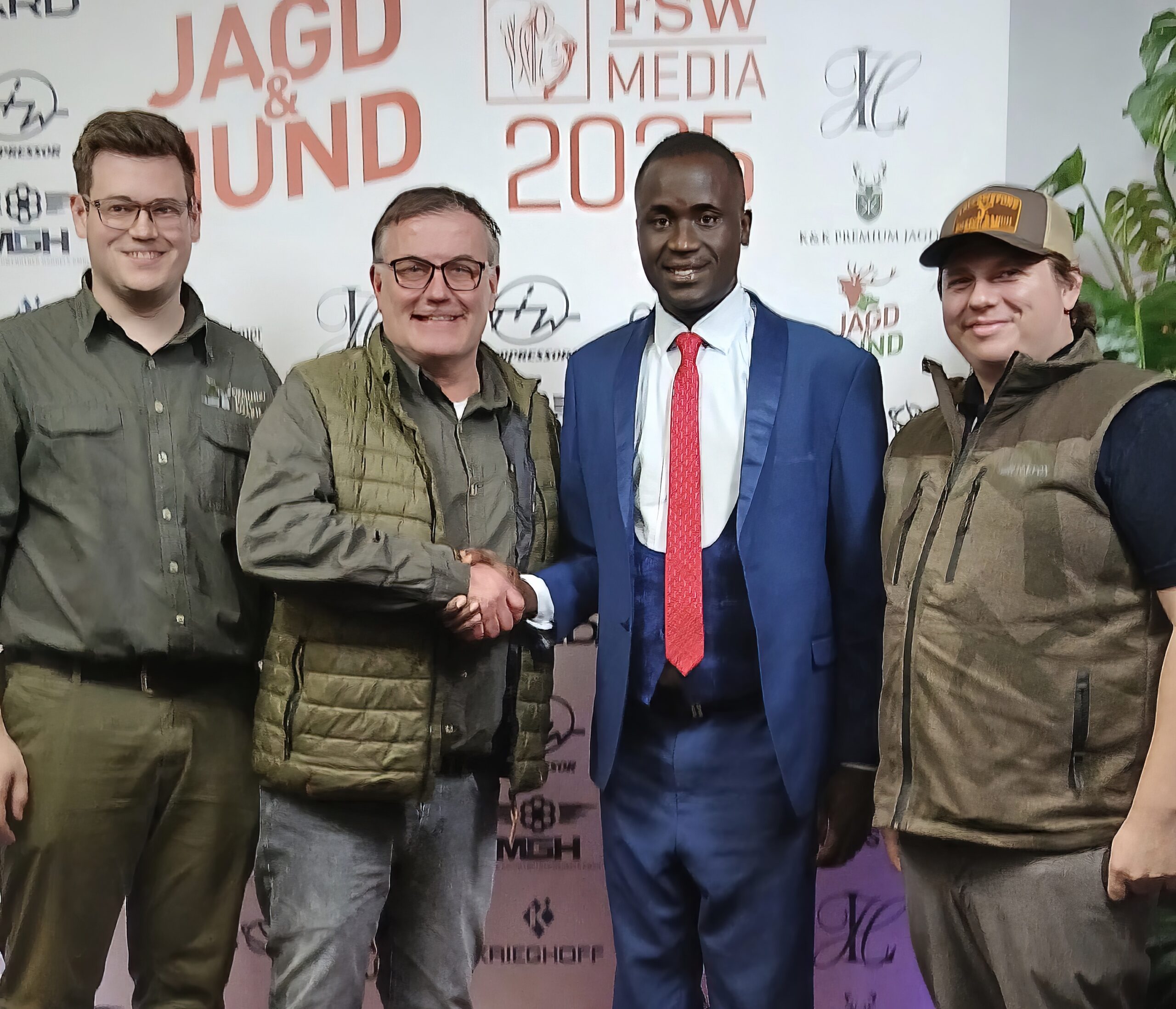 Two men shaking hands on stage whilst flanked by two others. One wears a smart suit, the others hunting greens.