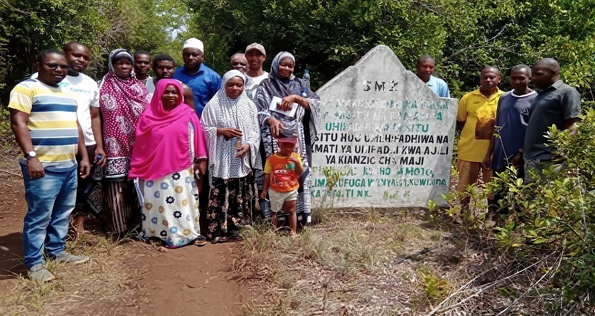A group of people posing with a stone sign painted with KiSwahili text.